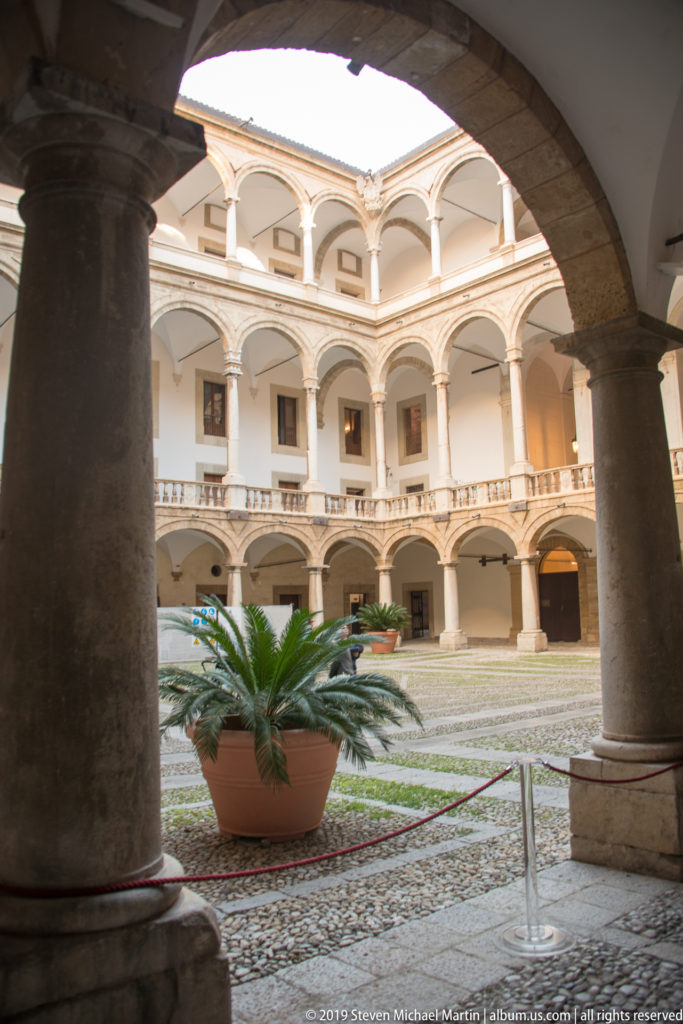 Sunlit two-story arcaded courtyard framed by a dark arch and column, with a large potted cycad in the center.