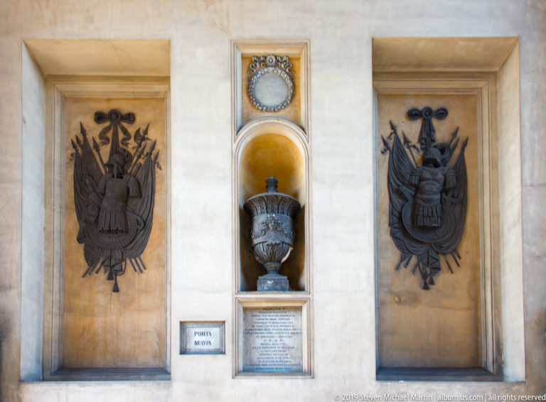 Two dark heraldic reliefs flank a central urn, circular emblem, and inscribed plaque set into a pale stone wall.