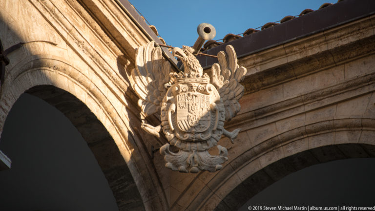 Sunlit crowned coat of arms with broad carved wings mounted high between two massive stone arches.