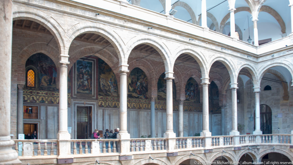 Upper courtyard gallery with repeated rounded arches, slender columns, balustrade, wall paintings, and visitors at the railing.