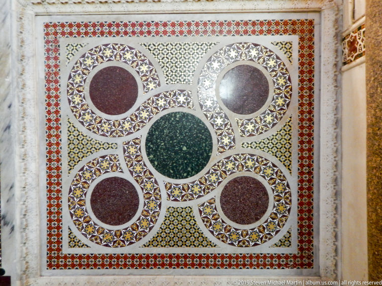 Square pavement panel with one green central disk and four reddish disks linked by multicolored geometric mosaic rings.