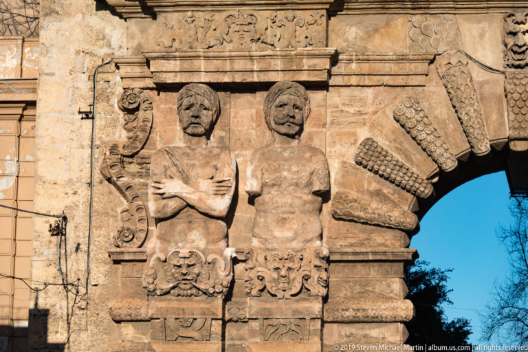 Two weathered male atlantes stand beside the radiating rusticated blocks of Porta Nuova’s main arch.