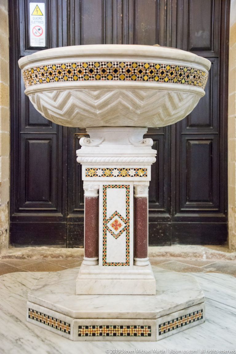 Inlaid marble basin on a pedestal before dark wooden doors.