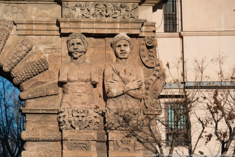 Close view of a bare-chested stone atlante with crossed arms beside masks and the rusticated arch of Porta Nuova.