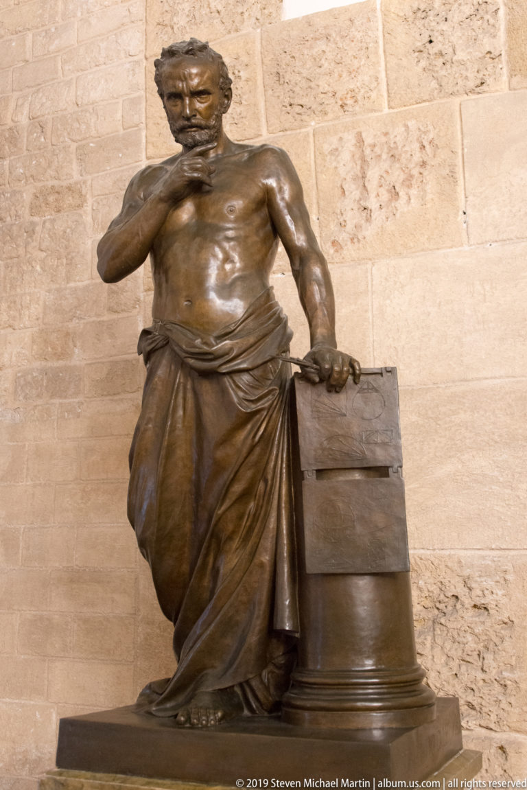 Bronze seated draped male figure beside a rectangular block.