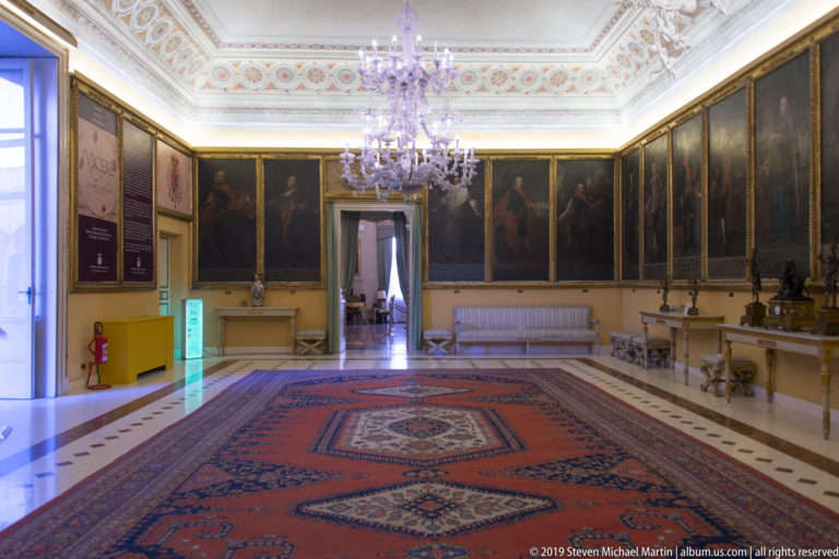 Formal palace portrait gallery with chandelier and patterned carpet.