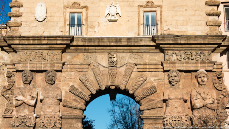 Two monumental male atlantes with crossed arms flank a rusticated stone arch decorated with masks and carved ornament.