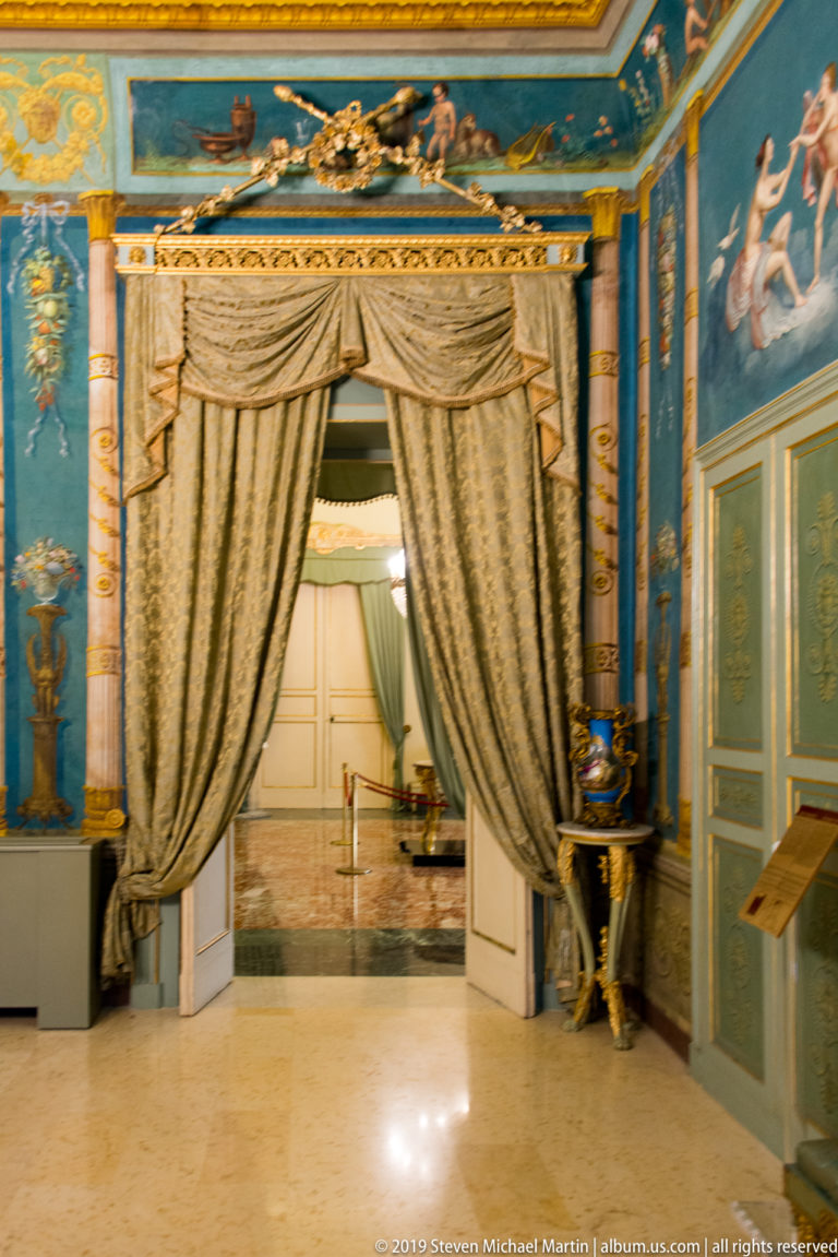 Blue and gold palace room with mythological wall paintings, classical pilasters, and heavy draperies framing an open doorway.