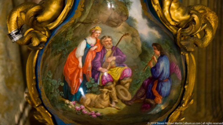 Small pastoral painting of three figures in a wooded setting enclosed by an elaborate gilded scrolling frame.