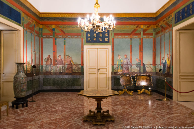 Formal palace room with chandelier, painted figural wall frieze, decorative cabinets, vase, central table, and marble floor.