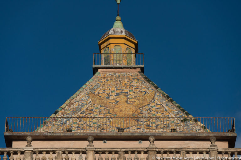 Pyramidal tiled roof with a giant eagle motif and small lantern above a stone balustrade under a blue sky.
