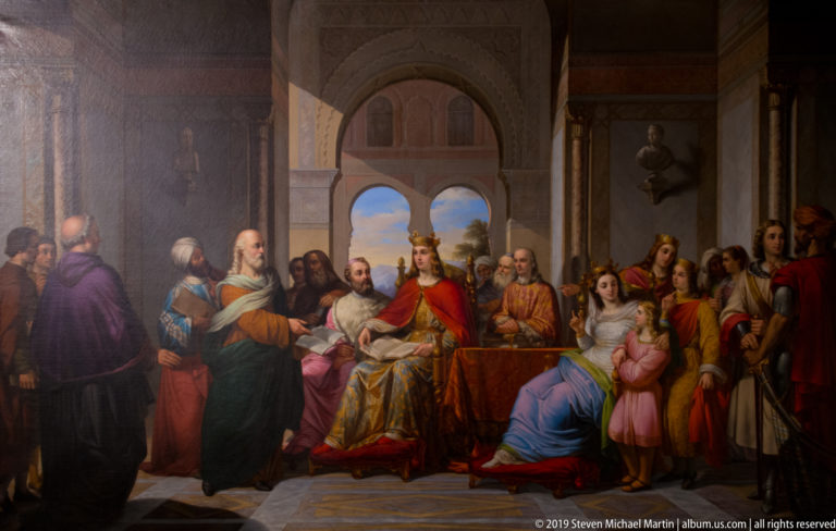Historical painting with a crowned ruler in red seated among clerics, courtiers, women, and children inside an arched hall.