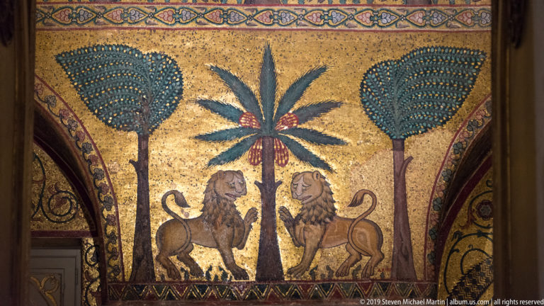 Two mosaic lions face a central date palm between tall stylized trees on a luminous gold background.