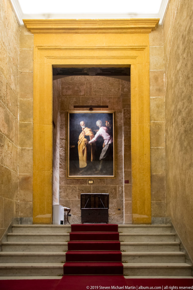 Framed painting of a reclining figure hangs above a red-carpeted stone staircase within a monumental palace opening.