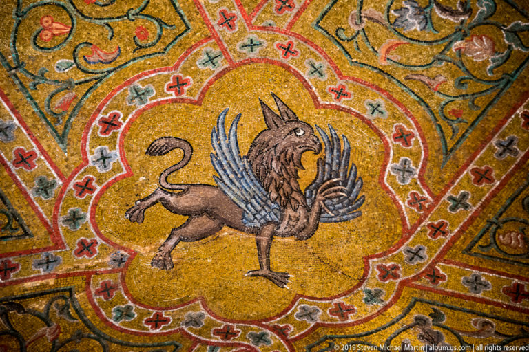 Winged lion in profile occupies a gold mosaic medallion surrounded by red, gray, and green geometric ornament.