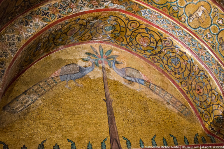 Two mosaic peacocks face a central stylized palm beneath a richly patterned arch on a gold background.