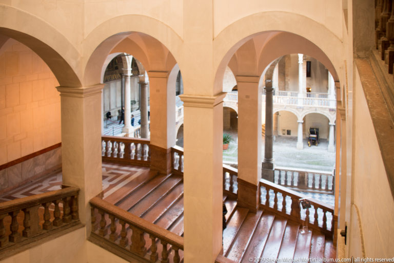 Three pale rounded arches frame a balustraded stair landing with the bright palace courtyard visible beyond.