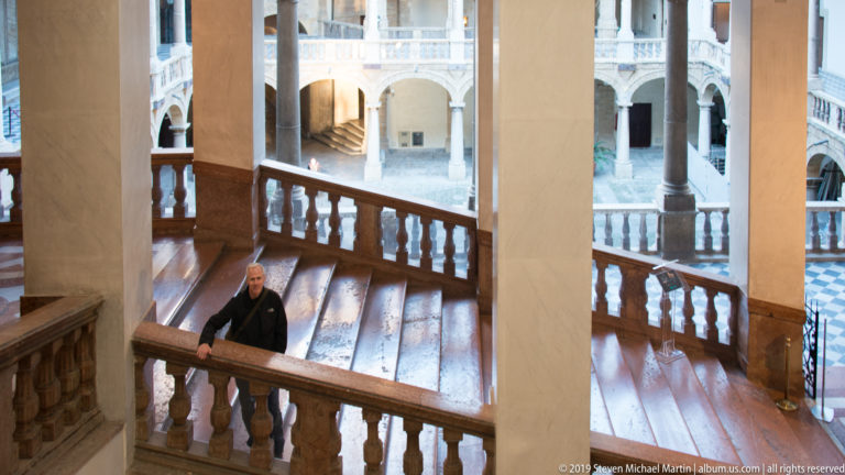 Visitor stands on a broad stone staircase with balustrades and tall piers overlooking the palace’s arcaded courtyard.