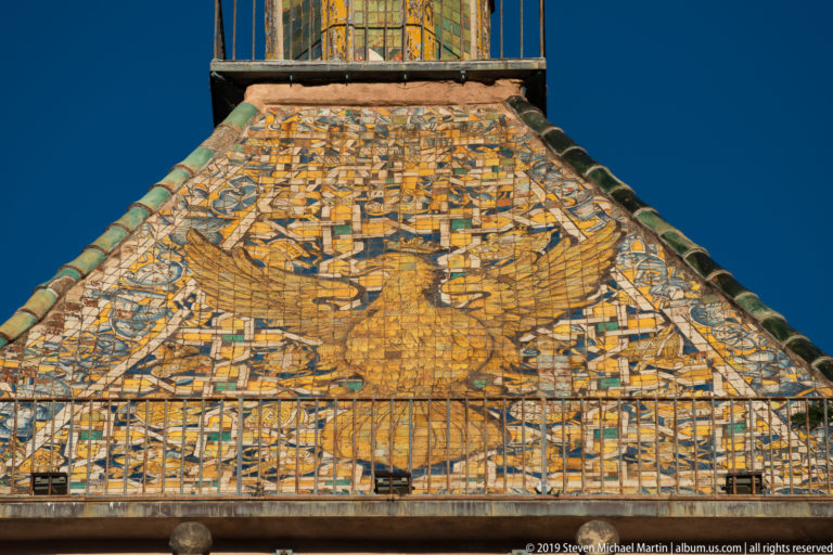 Large spread-winged eagle formed in colored roof tiles beneath a lantern, with a railing across the lower edge.