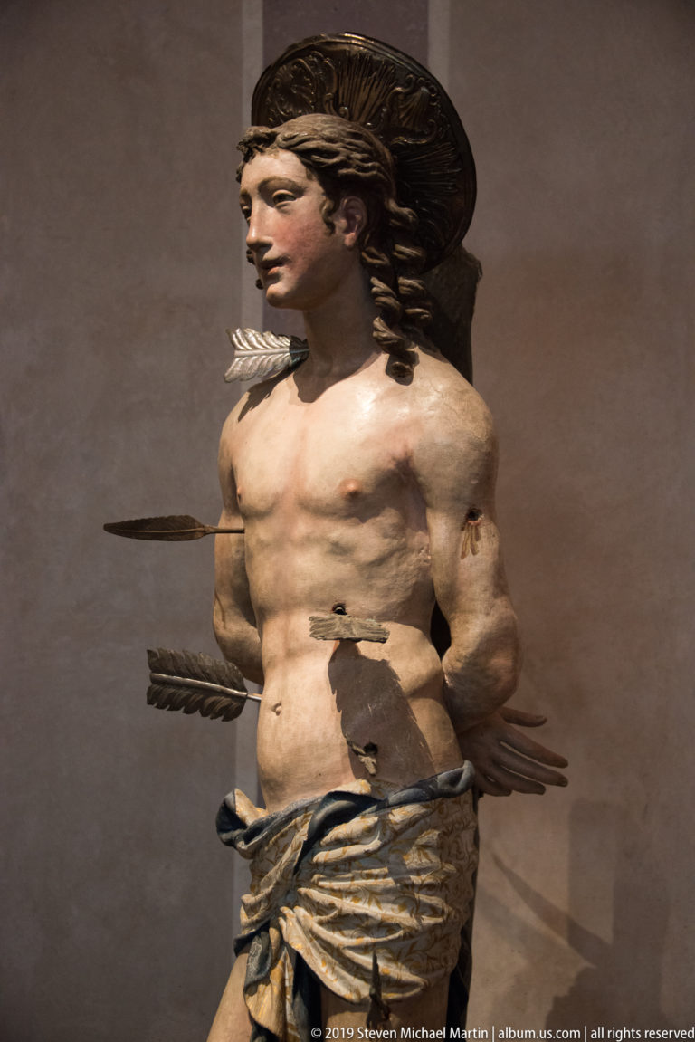 Standing sculpture of Saint Sebastian with bare torso, bound arms, arrows through his body, and drapery at his hips.