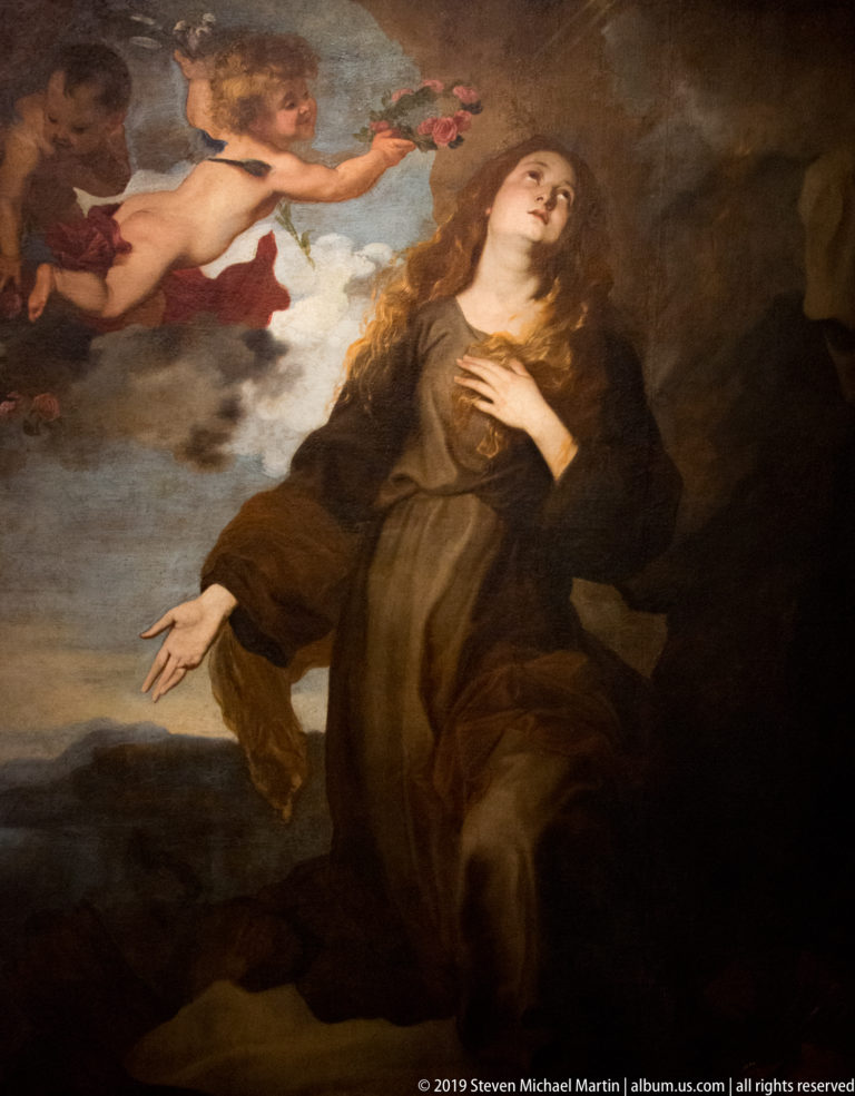 Female saint kneels in dark robes as an angel reaches toward her with flowers against a shadowed background.