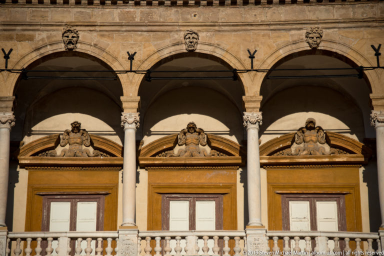 Three arched loggia bays with slender columns, balustrade, tall windows, and sculpted triangular pediments with reclining figures.