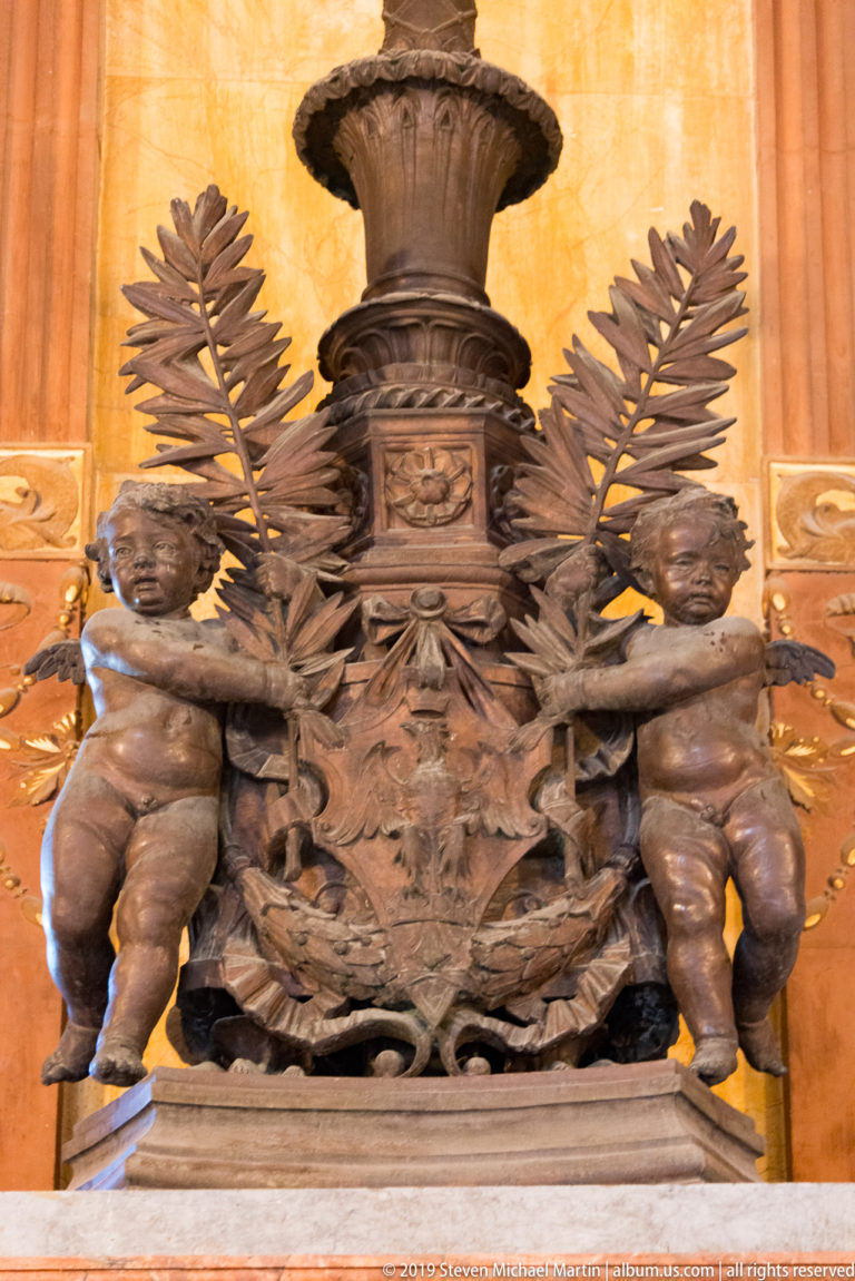 Dark ornamental urn above two seated putti, dense foliage, and fan-like fronds against warm stone.