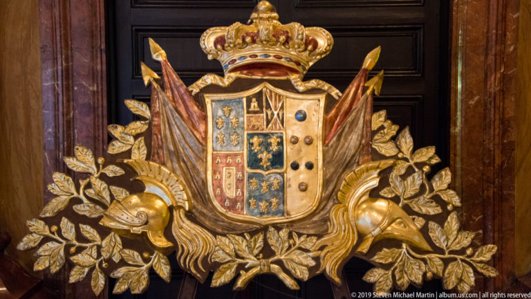 Crowned multicolored heraldic shield surrounded by gilded foliage, banners, and sculptural ornament.