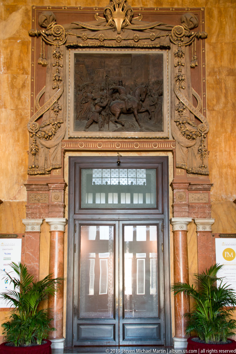 Dark double doors beneath a framed relief, flanked by reddish columns, carved ornament, and potted palms.