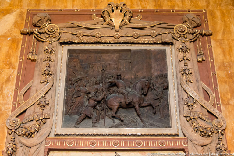 Dark equestrian relief with mounted figures inside an ornate frame of swags, ribbons, and rosettes.
