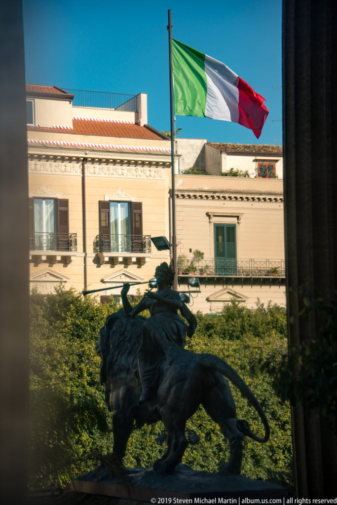 Italian flag flies above a dark bronze lion sculpture with a sunlit Palermo building beyond.