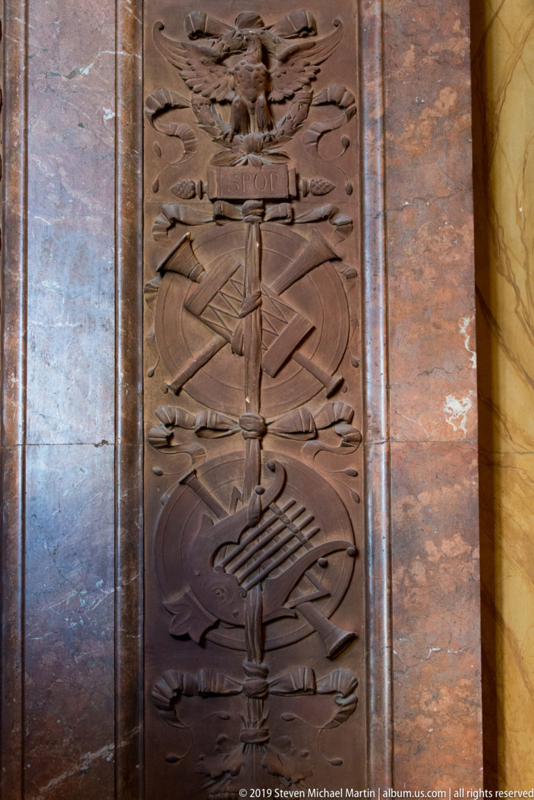 Full vertical carved trophy panel set between polished reddish-brown and pale stone wall surfaces.