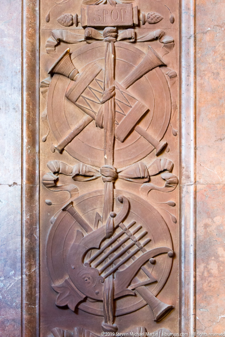 Vertical carved relief of crossed implements, ribbons, circular forms, and a lyre-like musical motif.
