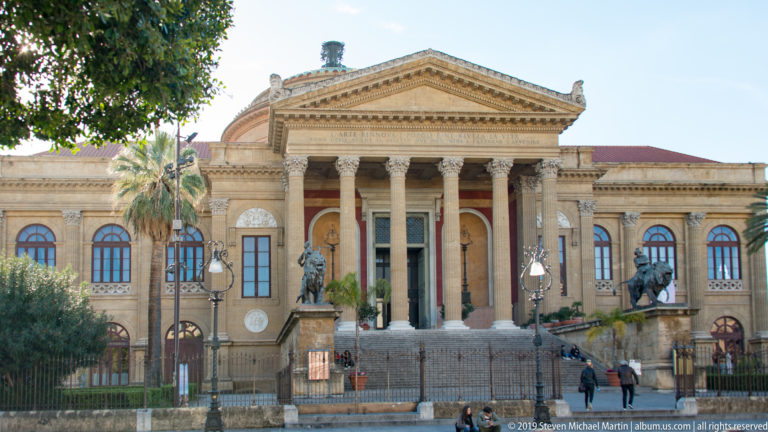 Wide view of the Teatro Massimo behind trees and fencing, with portico, wings, stairs, and bronze sculptures.