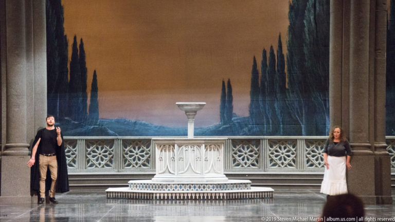 Two performers stand at opposite sides of a stage with cypress scenery and a pale central pedestal.