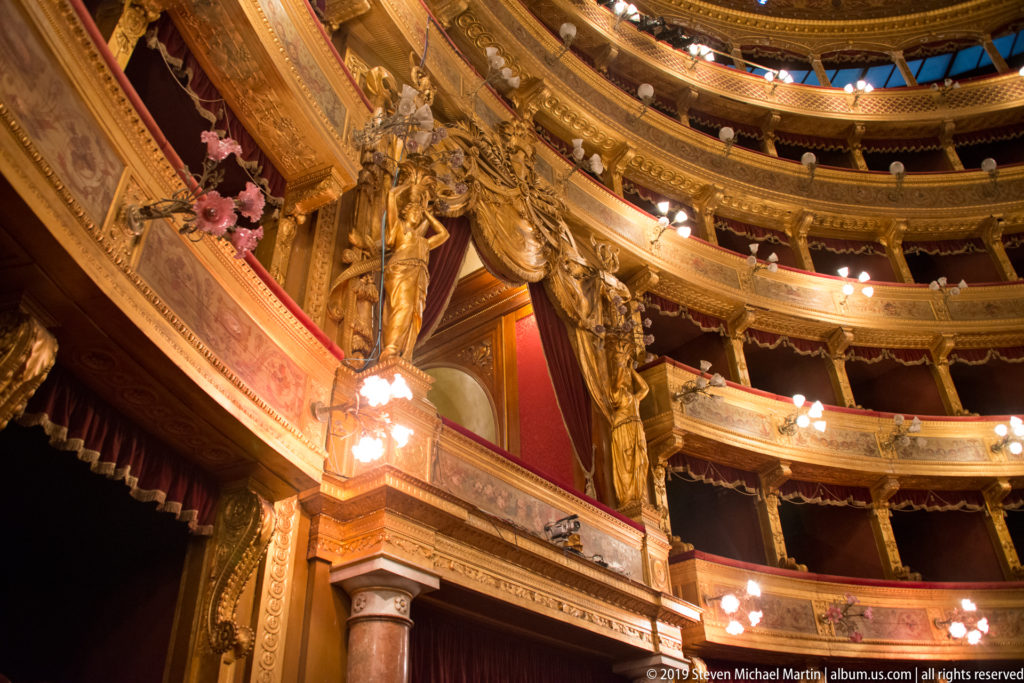 Curving tiers of illuminated gilded theater boxes rise beside sculptural figures at the stage opening.