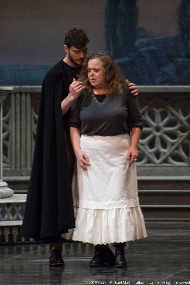 Two performers stand close together onstage, one in black and one in a long white skirt.