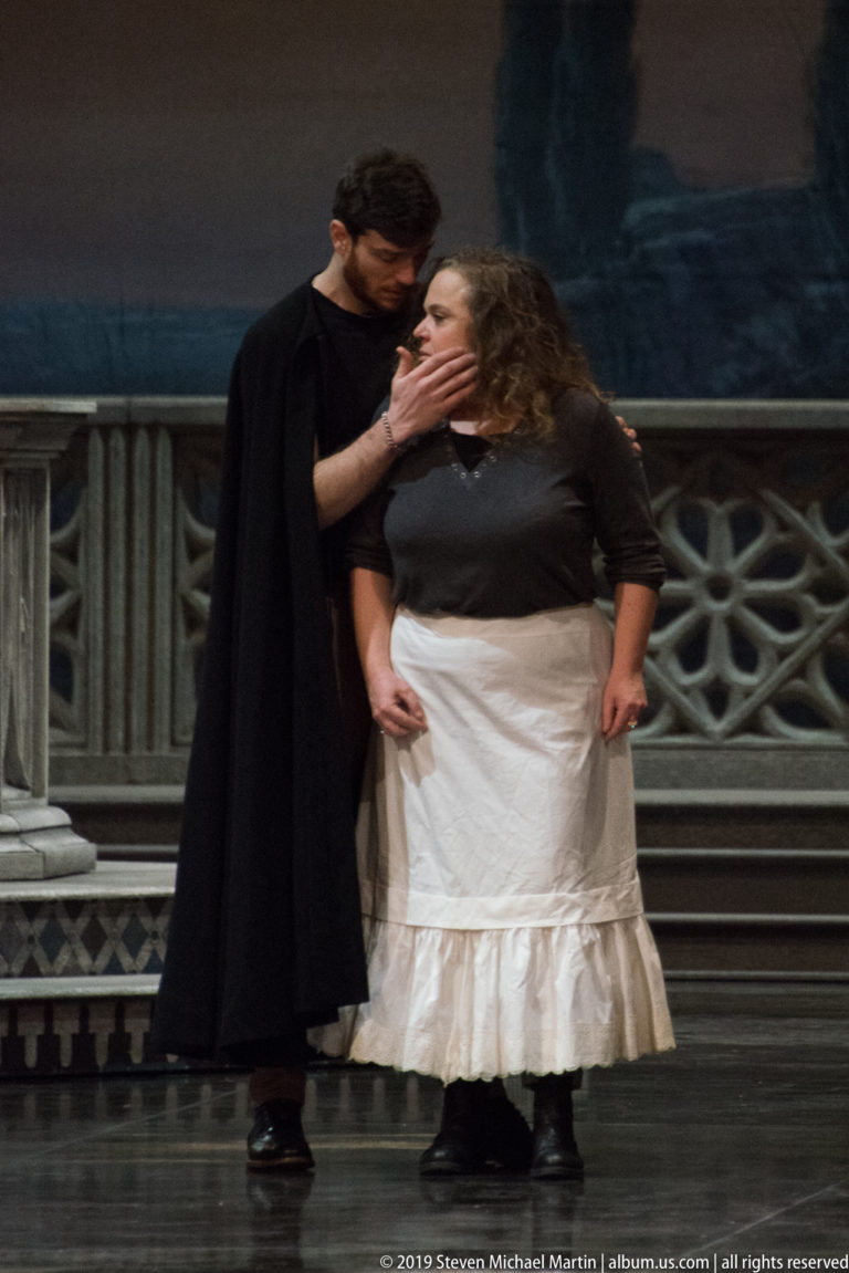 Black-clad performer reaches toward a woman in a white skirt before a painted stage backdrop.