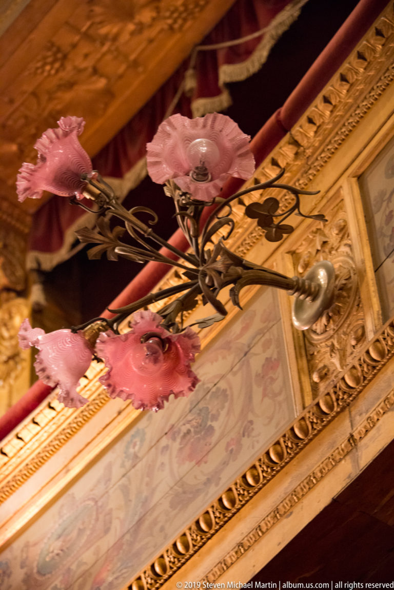 Dark scrolling light fixture with pink flower-shaped shades against gilded molding and red drapery.