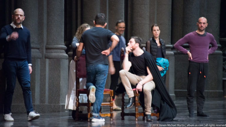 Performers in rehearsal clothing gather around a seated man onstage beside tall dark columns.