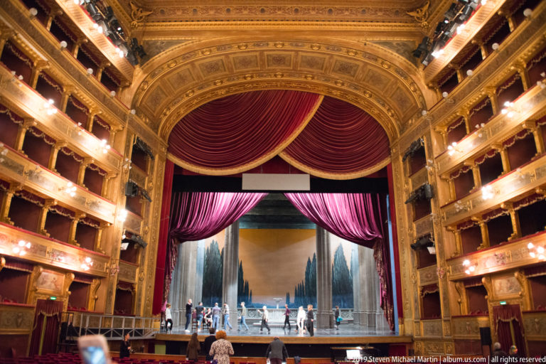 Centered auditorium view of an open stage framed by crimson curtains, ornate proscenium, illuminated box tiers, and several people.