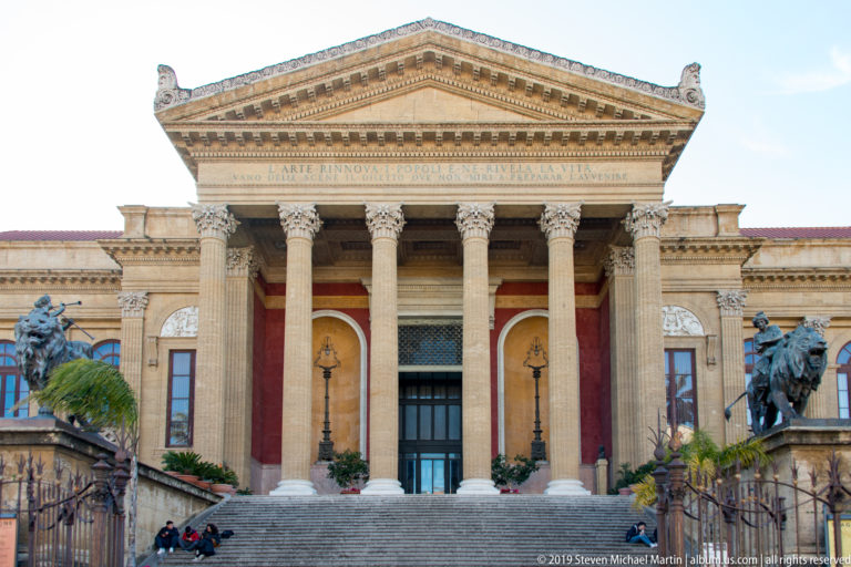 Centered Teatro Massimo façade with broad steps, columned portico, triangular pediment, and bronze lion sculptures.