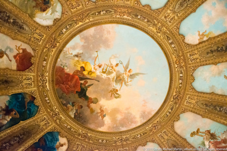 Ornate circular theater ceiling with a central sky painting, gilded borders, and surrounding painted panels.