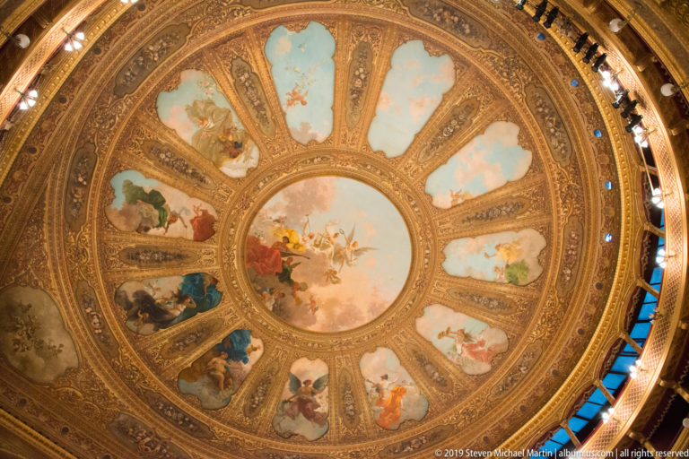 Large circular auditorium ceiling with a central painted scene and radiating figurative panels framed by elaborate gilded ornament.