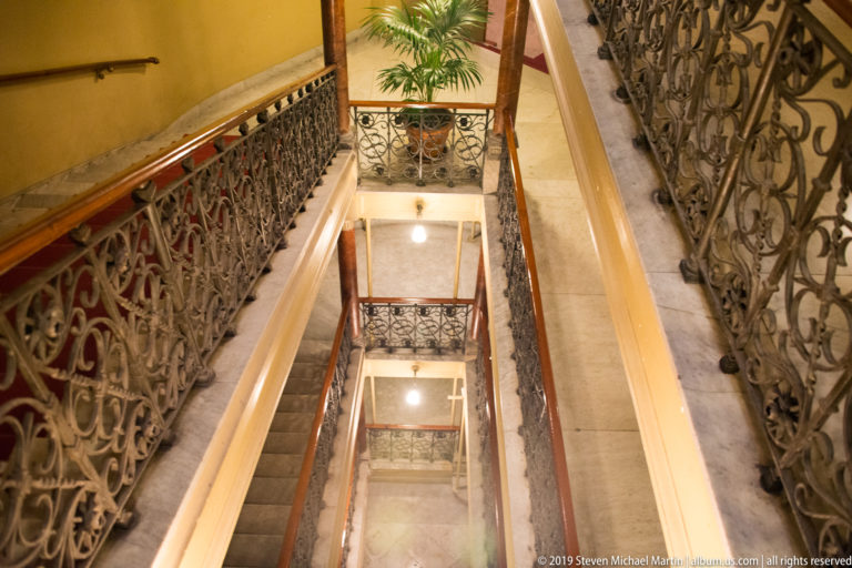 Downward view through several stairwell levels with ornate dark metal railings, wooden handrails, stone steps, and landings.