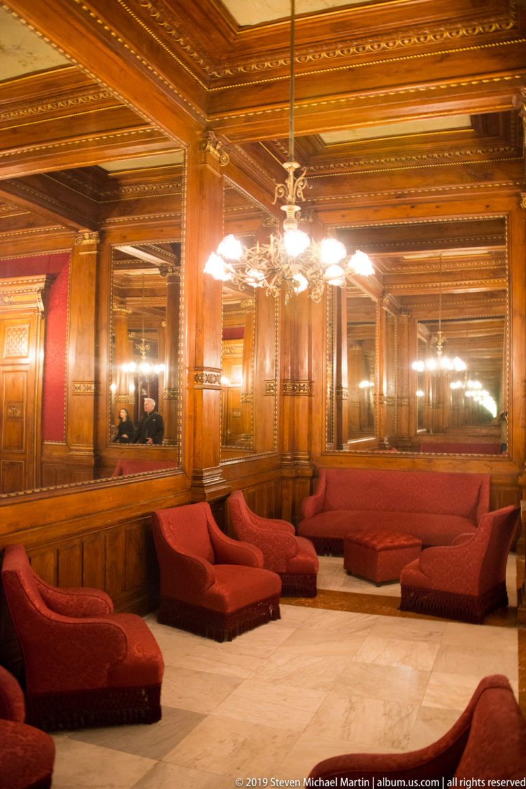 Red upholstered sofas and lounge seats arranged beneath chandeliers in a mirrored, polished wood-paneled reception salon.