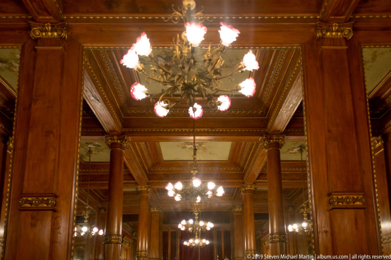 Large glowing chandelier hangs between polished wooden columns with mirrors and additional chandeliers receding behind it.
