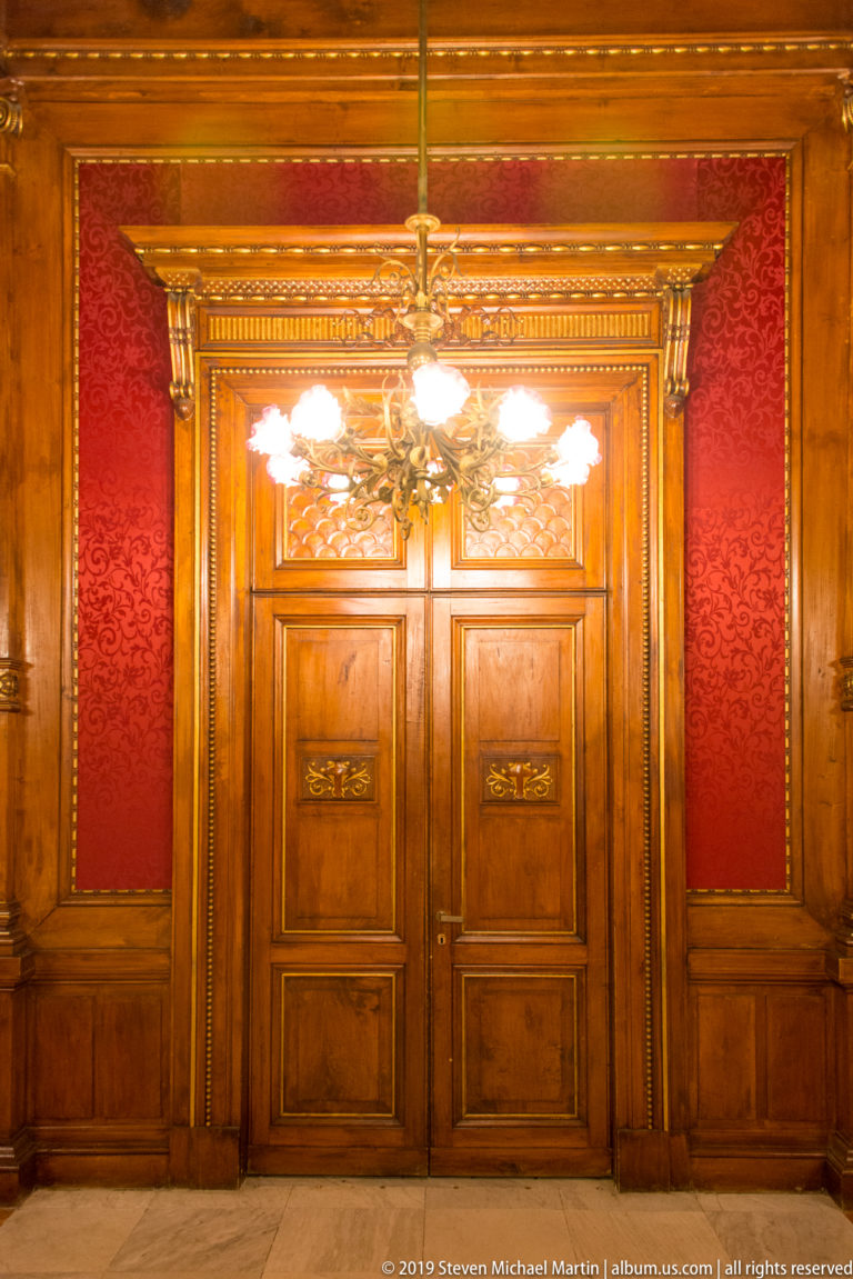 Tall carved wooden double doors centered between red damask wall panels beneath warm chandelier light.