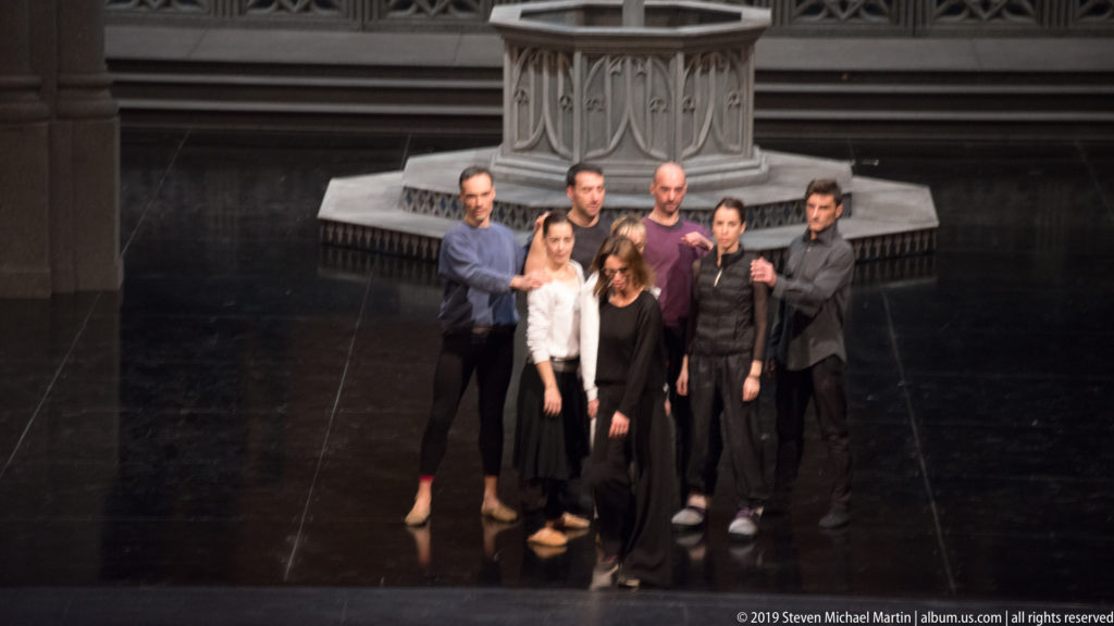 Eight performers in rehearsal clothing stand closely grouped on a dark theater stage beneath a large architectural structure.
