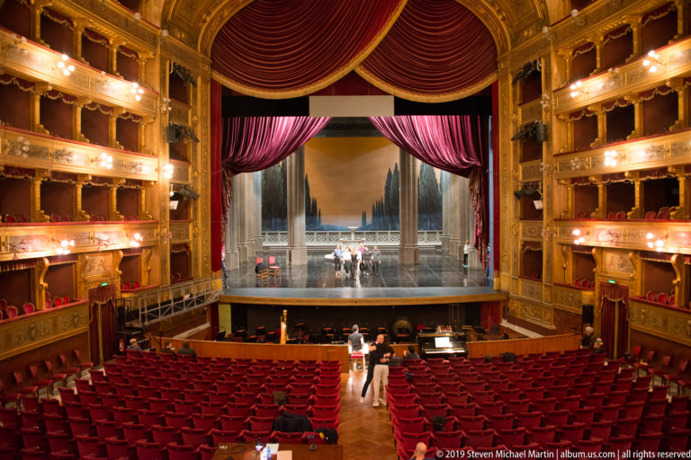 Wide view over red auditorium seats toward an open stage with people and equipment, flanked by illuminated gilded box tiers.